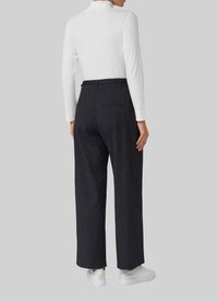 Personne portant un pantalon large à rayures noires, un haut à manches longues blanc à côtes, et des baskets blanches, se tenant contre un fond uni.