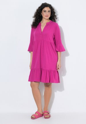 Kvinde med krøllet hår iført en knælængde magenta kjole med flæsekant og 3/4 ærmer, kombineret med matchende magenta sandaler.