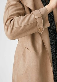 Beżowy płaszcz z zamszu z rękawem ozdobionym klamrą, z widoczną haftowaną kieszenią oraz częścią czarnej sukienki w wzory pod spodem.