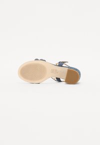 Denim hælhæl sandal med en beige sål, der har to stropper og en spændeankelrem. Tekstureret gummisål med ribbet mønster.
