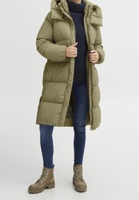 Lang olivengrøn pufferjakke med hætte, der har en quiltet design, store lommer og lynlåslukning. Båret med blå jeans og brune støvler.