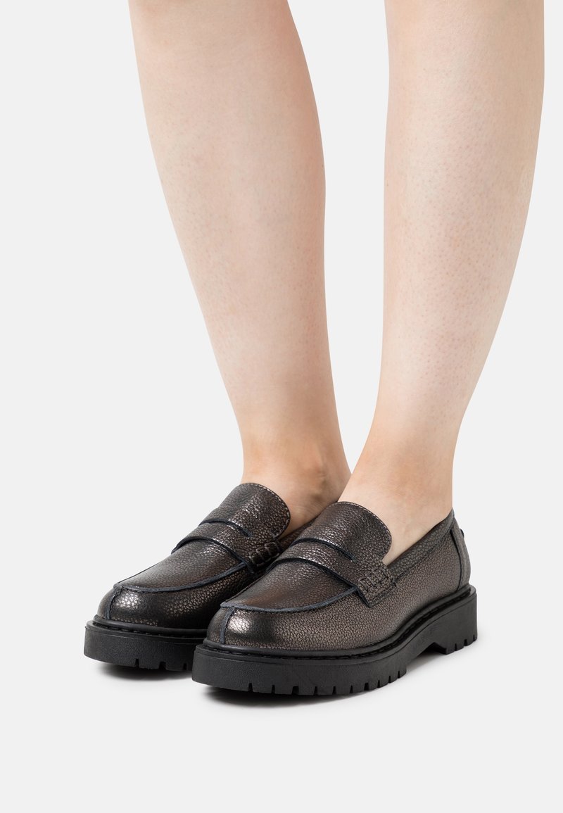 Mocassins en cuir marron foncé brillant avec des semelles épaisses noires à relief portés pieds nus sur un fond clair uni.