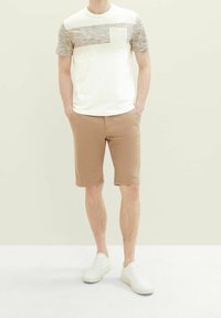 Homme portant un short beige, des baskets blanches et un t-shirt blanc avec une bande horizontale multicolore et une poche poitrine.