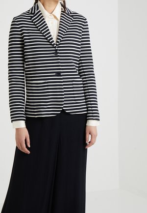 Vrouw die een blazer met horizontale marineblauwe en witte strepen draagt over een wit overhemd met kraag en zwarte broek met wijde pijpen.