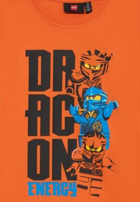 Oranž T-särk, millel on musta paksu tekstiga "DRAGON ENERGY", ja kolm LEGO Ninjago figuuri sinises ja oranžis, esitledes keerukaid disaine.