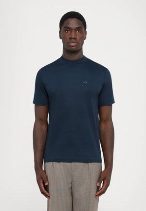 Young man wearing a navy blue short-sleeve shirt and light gray pants, standing against a plain white background.