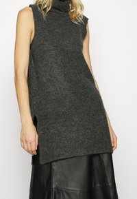 Tunique sans manches à col roulé gris foncé avec une texture côtelée, des fentes latérales et une coupe décontractée, associée à une jupe en similicuir noire.