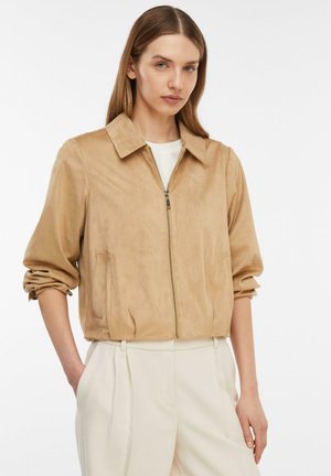 Kobieta ubrana w beżową kurtkę na zamek z kołnierzem i marszczonymi rękawami, w połączeniu z białymi plisowanymi spodniami z wysokim stanem, stojąca z rękami w kieszeniach.