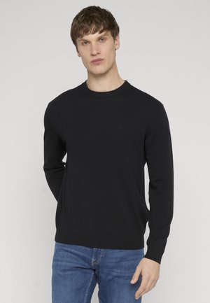 Mężczyzna o krótkich brązowych włosach, ubrany w czarny sweter z długim rękawem i niebieskie dżinsy, stojący na jasnym jednolitym tle.