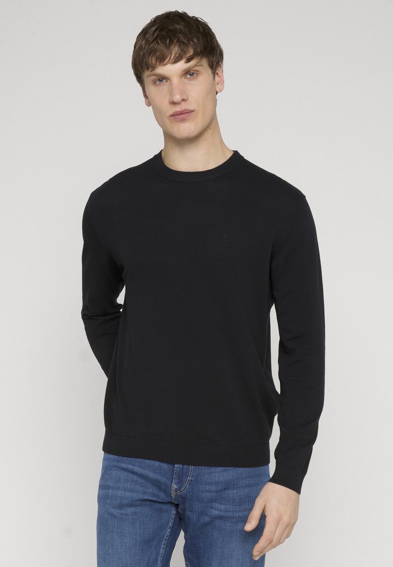 Homme aux cheveux bruns courts portant un pull noir à manches longues et un jean bleu, debout devant un fond clair uni.