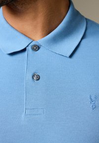 Světle modrá polo košile s texturovanou látkou, disponující límcem, dvěma šedými knoflíky a malým vyšitým logem jelena na prsou.