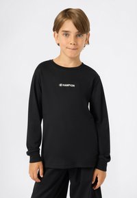 Jeune personne portant un t-shirt à manches longues noir avec le logo "Champion", se tenant contre un fond blanc, les mains détendues à ses côtés.