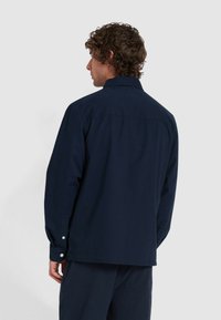 Mężczyzna z kręconymi włosami, w ciemnoniebieskiej koszuli z długim rękawem oraz strukturalnych niebieskich spodniach, stoi i jest odwrócony plecami do jednolitego tła.