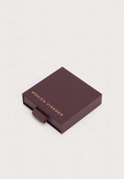 Σκούρο καφέ τετράγωνο κουτί κοσμημάτων με υφασμάτινη θηλιά τραβήγματος και χρυσά γράμματα στην καπάκι που γράφουν "MONICA VINADER".