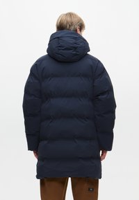 Námořnická modrá péřová bunda s prostornou kapucí, prošívaným designem a elastickými manžetami, sahající do poloviny stehna s minimalistickou estetikou.