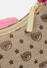 Plátěná taška Tan s motivy hnědých hvězd a rukou, růžovým vnitřkem, zlatým zipem a vyrytým štítkem s názvem značky.