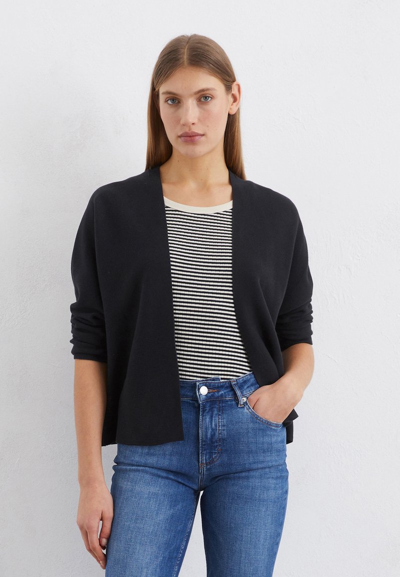 Schwarze, offenherzige Strickjacke aus weichem Material mit langen Ärmeln. Kombiniert mit einem schwarz-weißen gestreiften Shirt und blauen Jeans.
