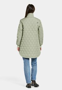 Quiltad, lång olivgrön täckjacka med ståkrage, som har diamantmönstrade detaljer och ribbade mudd. Bärs över blå jeans och svarta skor.