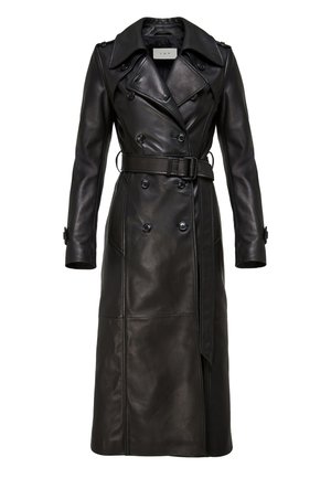 Woman standing wearing a long black leather belted trench coat, black pointed boots, holding a large black fuzzy tote bag.