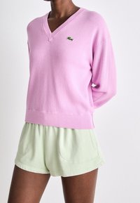 Suéter de pico en rosa claro de tejido suave con dobladillo acanalado, combinado con pantalones cortos verdes claros que tienen bolsillos frontales y una cinturilla elástica.