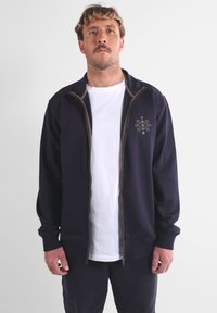 Veste zippée bleu marine au design ajusté, texture douce, fermeture éclair marron contrastante et un subtil motif de flocon de neige brodé sur la poitrine.