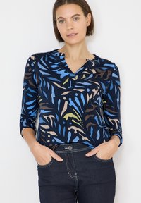 Granatowy top z żywym abstrakcyjnym wzorem w odcieniach niebieskiego, zielonego i brązowego. Posiada dekolt henley oraz rękawy do łokcia.