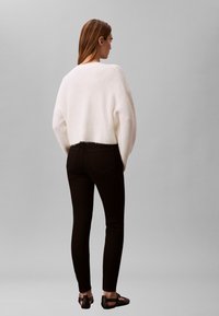 Balts pūkains džemperis, tievās melnās džinsu bikses un melnas plakanas kurpes. Mīksta tekstūra, brīvs piegriezums ar nolaižamām plecu līnijām un saīsinātu garumu mugurpusē.