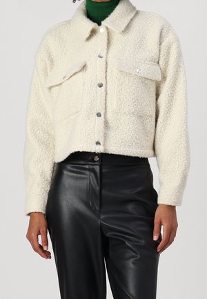 Person trägt eine cremefarbene, strukturierte Jacke mit Knopfleiste und zwei Patentaschen, kombiniert mit schwarzen, hoch taillierten Lederhosen.