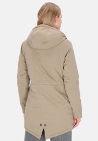 Kvinde iført en beige hætteparka med justerbar talje og rygventilation, stående med ryggen mod en ensfarvet baggrund.