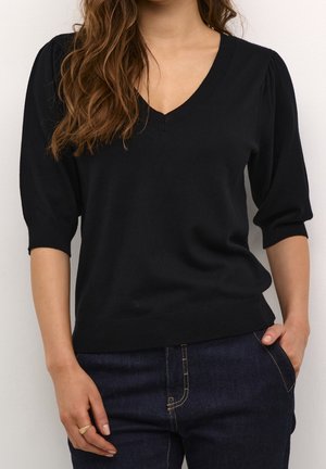 Kvinde iført en sort V-hals kort puffet ærmet sweater og mørkeblå jeans med den ene hånd i lommen og den anden hvilende ved siden af sig.