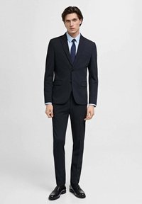 Costum de marine cu două piese, format dintr-o jachetă croită și pantaloni slim-fit. Detalii notabile includ o cămașă albastră deschis și o cravată închisă. Pantofi din piele neagră.