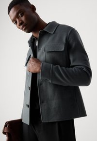 Gray wool blend jacket with a button front, two chest pockets, and a collar, worn by a man holding a brown leather bag.