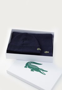 Tamsiai mėlynos megztos kepurės ir šaliko rinkinys su žaliais krokodilo logotipais, pateiktas baltame dėžutėje su žaliu krokodilu ant dangčio.