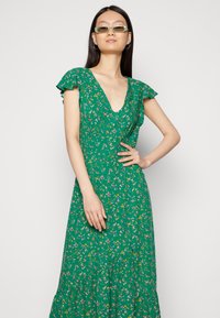 Vestido floral verde com mangas curtas e esvoaçantes, decote em V profundo e saia em camadas, apresentando pequenos padrões de flores multicoloridas por toda parte.