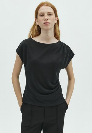 Junge Frau mit roten Haaren, die ein schwarzes Kurzarm-Top und schwarze Hosen trägt, steht mit den Händen in den Taschen vor einem einfarbigen Hintergrund.