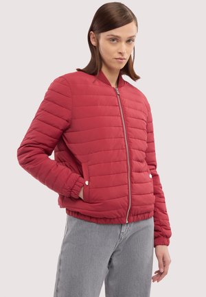 Kobieta o prostych brązowych włosach, ubrana w czerwoną pikowaną kurtkę zapinaną na zamek i szare dżinsy, stojąca z jedną ręką w kieszeni.