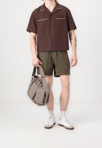 Brązowa koszula z krótkim rękawem zapinana na guziki z białymi lamówkami, oliwkowe szorty, szara materiałowa torba na ramię, białe sneakersy z brązowymi akcentami.