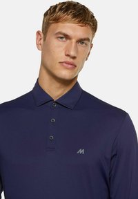 Junger Mann mit kurzem blonden Haar, der ein dunkelblaues langärmliges Poloshirt mit drei Knöpfen und einem kleinen "M"-Logo auf der Brust trägt.