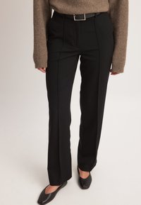 Čierne konferenčné nohavice s vysokým pásom, prednými záhybmi, hladkým povrchom a striebornou obdĺžnikovou prackou na opasku.