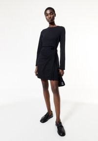 Zwarte jurk met lange mouwen, wikkelontwerp en strikkraag bij de taille, gecombineerd met zwarte loafers. Model met een kleine zwarte tas.