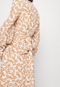 Weiße Bluse aus Stoff mit einem bedruckten braunen Blattmuster, langen Ärmeln und einem gebundenen Taillen-Detail, weicher Textur und ausgestelltem Design.