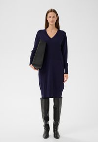 Marineblaues Strickkleid mit V-Ausschnitt und langen Ärmeln. Kombiniert mit einer rechteckigen schwarzen Handtasche und schwarzen Lederstiefeln über dem Knie.