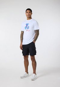 Muž s tetovanými pažemi a nohama, který má na sobě bílé tričko s grafikou, černé cargo šortky a bílé tenisky, stojící na jednobarevném pozadí.