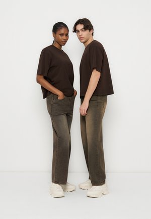 To voksne, vendt mod fronten, iført matchende mørkebrune løstsiddende t-shirts, falmede brune jeans med vide ben og hvide sneakers, stående mod en hvid baggrund.