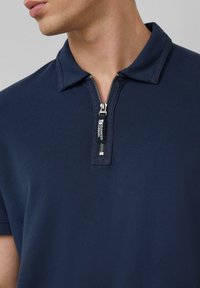 Granatowa koszulka polo o teksturowanym splocie, z suwakiem przy kołnierzyku oraz kontrastującym uchwytem suwaka. Krótkie rękawy i klasyczny krój.