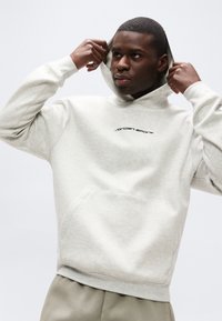 Νέος άνδρας που ρυθμίζει γκρι ανοιχτό hoodie με το λογότυπο "Jordan Sports", φοράει μπεζ παντελόνι μπροστά σε απλό λευκό φόντο.