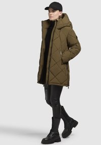 Olivgrüne gesteppten Pufferjacke mit Kapuze, Reißverschluss und Seitentaschen. Kombiniert mit schwarzen Lederleggings und geschnürten schwarzen Combat-Boots.