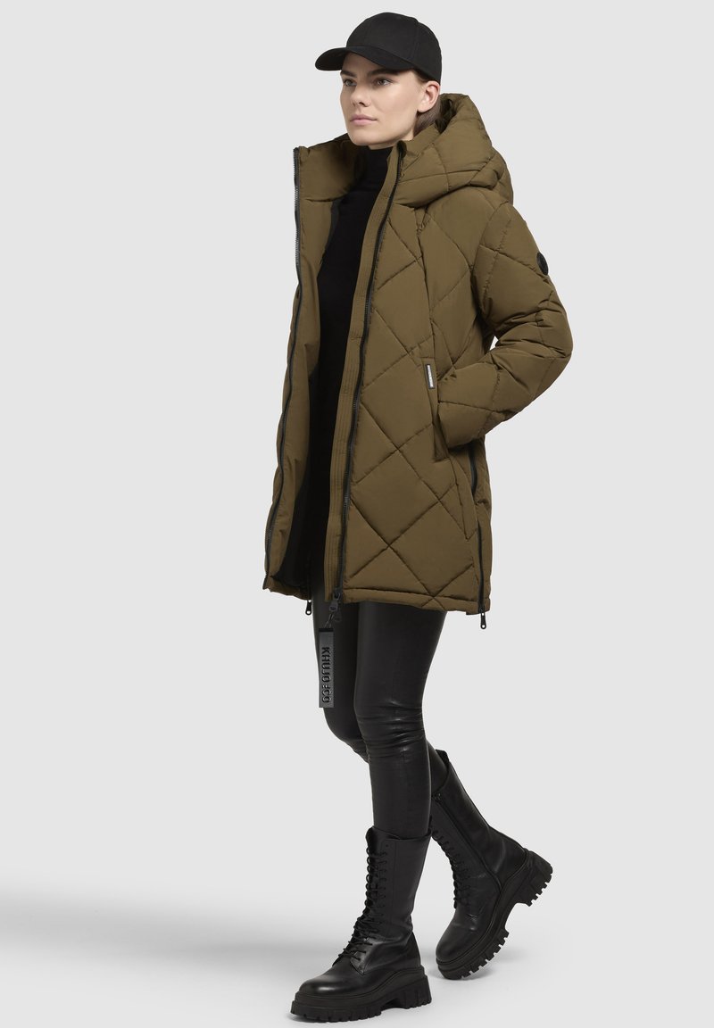 Cappotto imbottito trapuntato verde oliva con cappuccio, chiusura con zip e tasche laterali. Abbinato a leggings in pelle nera e stivali combat neri con lacci.