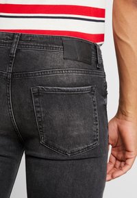 Jeans de denim negros de corte ajustado con una textura suave, que cuentan con dos bolsillos traseros y un parche de cuero en la cintura. Camisa de rayas horizontales visible arriba.