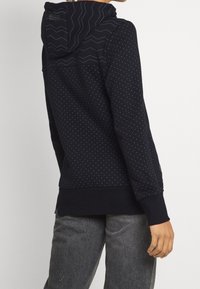 Sudadera azul marino con patrón texturizado, que incluye una capucha en zigzag, lunares y puños acanalados. Tela suave y corte relajado.